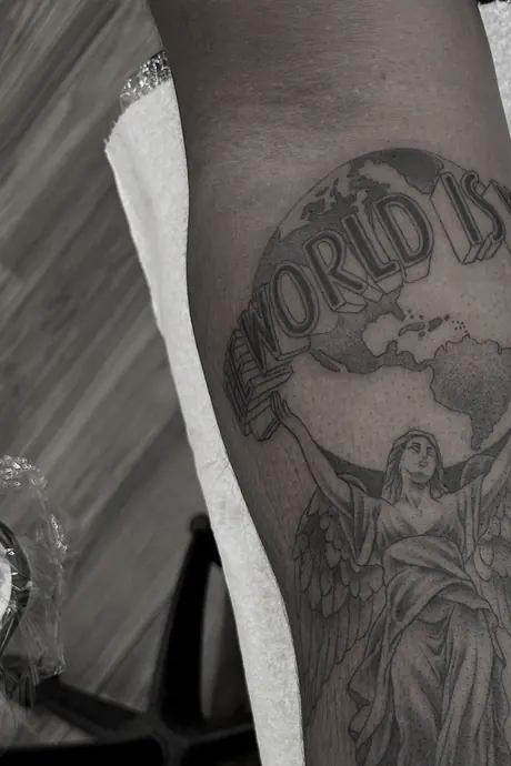 Black and grey forearm piece of a robed figure lifting a globe, with banner lettering reading The World Is across it.